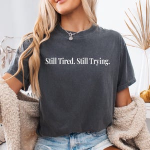 May include: Dark gray t-shirt with the text "Still Tired. Still Trying." in white. The shirt has a crew neck and short sleeves. The model is wearing a silver necklace and a cream-colored cardigan.