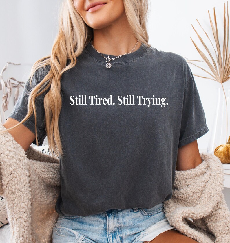 May include: Dark gray t-shirt with the text "Still Tired. Still Trying." in white. The shirt has a crew neck and short sleeves. The model is wearing a silver necklace and a cream-colored cardigan.