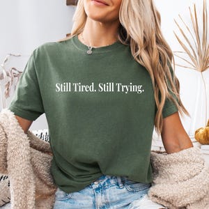 May include: Olive green t-shirt with the text "Still Tired. Still Trying." in white. The shirt is a short-sleeved, crew neck style. The model is wearing blue denim shorts and a beige cardigan.