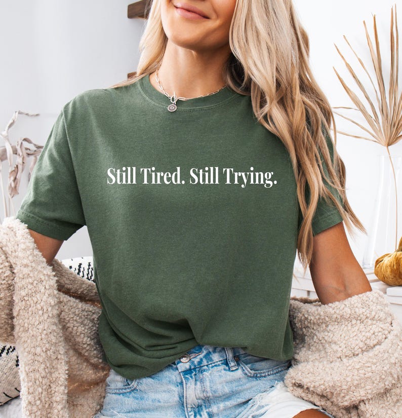 May include: Olive green t-shirt with the text "Still Tired. Still Trying." in white. The shirt is a short-sleeved, crew neck style. The model is wearing blue denim shorts and a beige cardigan.