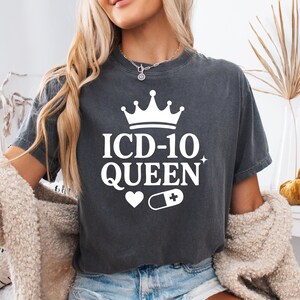 May include: Dark gray t-shirt with the text "ICD-10 QUEEN" in white, featuring a crown, heart, and pill graphic. The shirt is a crew neck style and appears to be made of a soft material.
