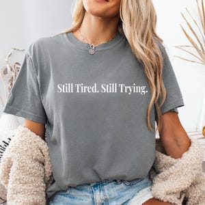 May include: A gray t-shirt with the text "Still Tired. Still Trying." in white. The shirt is a crew neck style and appears to be made of a soft material. The person in the image is wearing a necklace and a cream-colored sweater.