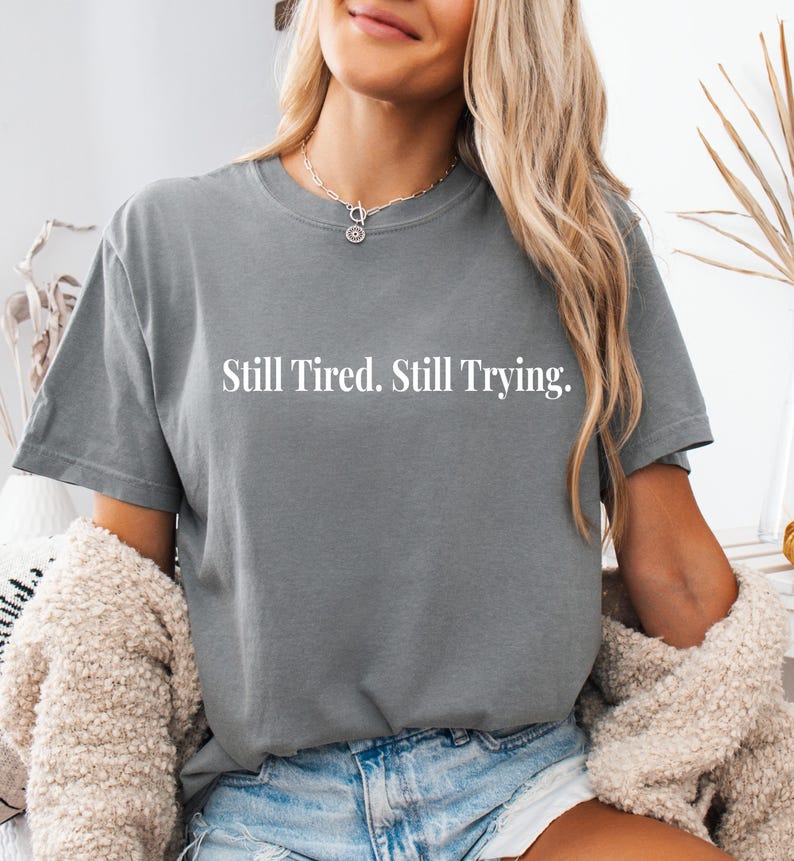 May include: A gray t-shirt with the text "Still Tired. Still Trying." in white. The shirt is a crew neck style and appears to be made of a soft material. The person in the image is wearing a necklace and a cream-colored sweater.
