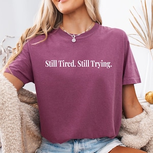 May include: A short-sleeved, maroon t-shirt with the text "Still Tired. Still Trying." in white block letters. The shirt is made of a soft material and has a relaxed fit. The shirt is a casual wear item.