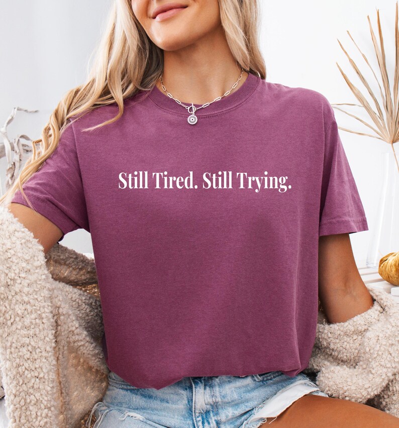 May include: A short-sleeved, maroon t-shirt with the text "Still Tired. Still Trying." in white block letters. The shirt is made of a soft material and has a relaxed fit. The shirt is a casual wear item.