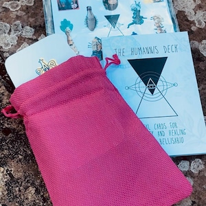Peut inclure: Un sac rose à cordon est posé devant deux jeux de cartes. L'un est intitulé "ART DECK" et l'autre "THE HUMANIS DECK". Les cartes présentent des illustrations et du texte. L'arrière-plan est une surface naturelle texturée.