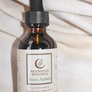 Puede incluir: Una botella de vidrio marrón con un cuentagotas negro, etiquetada como "MOONFOLK BOTANICA Aura Armor". La etiqueta incluye texto sobre un tónico protector. La botella está sobre un fondo blanco texturizado.