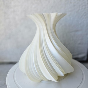 May include: A cream-colored, abstract vase with a swirling, layered design. The vase has a wide base that tapers to a narrow neck and flares out at the top. The design is made of many curved, vertical elements.