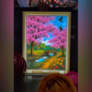 May include: A framed landscape art piece featuring a serene scene with pink flowering trees, a bridge over a stream, and birds in a blue sky. The artwork is illuminated by a soft, warm light, creating a tranquil ambiance. Decorative flowers are in the foreground.