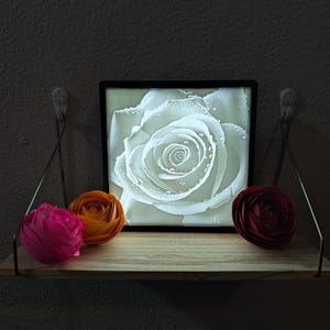 May include: A wooden shelf displays a framed white rose image and three paper flowers. The flowers are pink, orange, and burgundy. The framed image is a close-up of a white rose with water droplets.