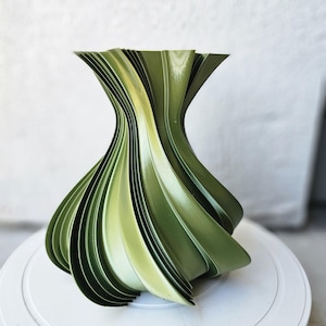 May include: A modern, abstract vase in shades of green. The vase has a unique, layered design with curved, leaf-like shapes. The glossy finish reflects light, highlighting the form's intricate details. The vase sits on a white surface.