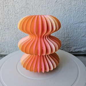 Puede incluir: Un jarrón decorativo abstracto y en capas en tonos naranja y rosa. El jarrón presenta una estructura apilada distintiva con una textura acanalada. El degradado de color cambia de naranja brillante en la parte superior a rosa suave en la base, creando una pieza visualmente atractiva.