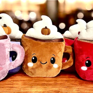 May include: A collection of plush coffee mugs in pink, brown, and red, each topped with whipped cream. The mugs have smiling faces and decorative elements like a snowflake, pumpkin, and heart. They are arranged on a wooden surface.