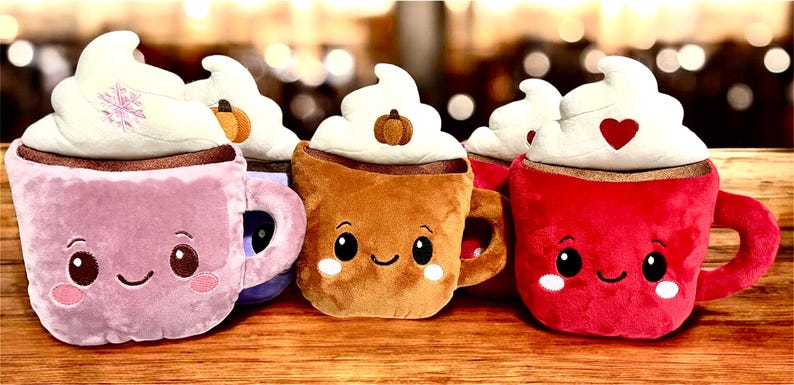 May include: A collection of plush coffee mugs in pink, brown, and red, each topped with whipped cream. The mugs have smiling faces and decorative elements like a snowflake, pumpkin, and heart. They are arranged on a wooden surface.