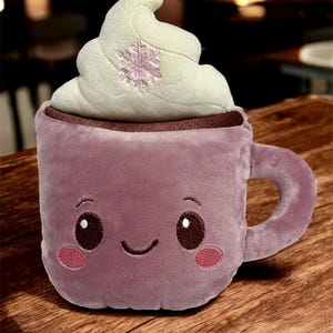 May include: Plush purple mug with a smiling face, pink cheeks, and a whipped cream topping. The whipped cream has a pink snowflake embroidered on it. The mug has a brown rim and a handle.