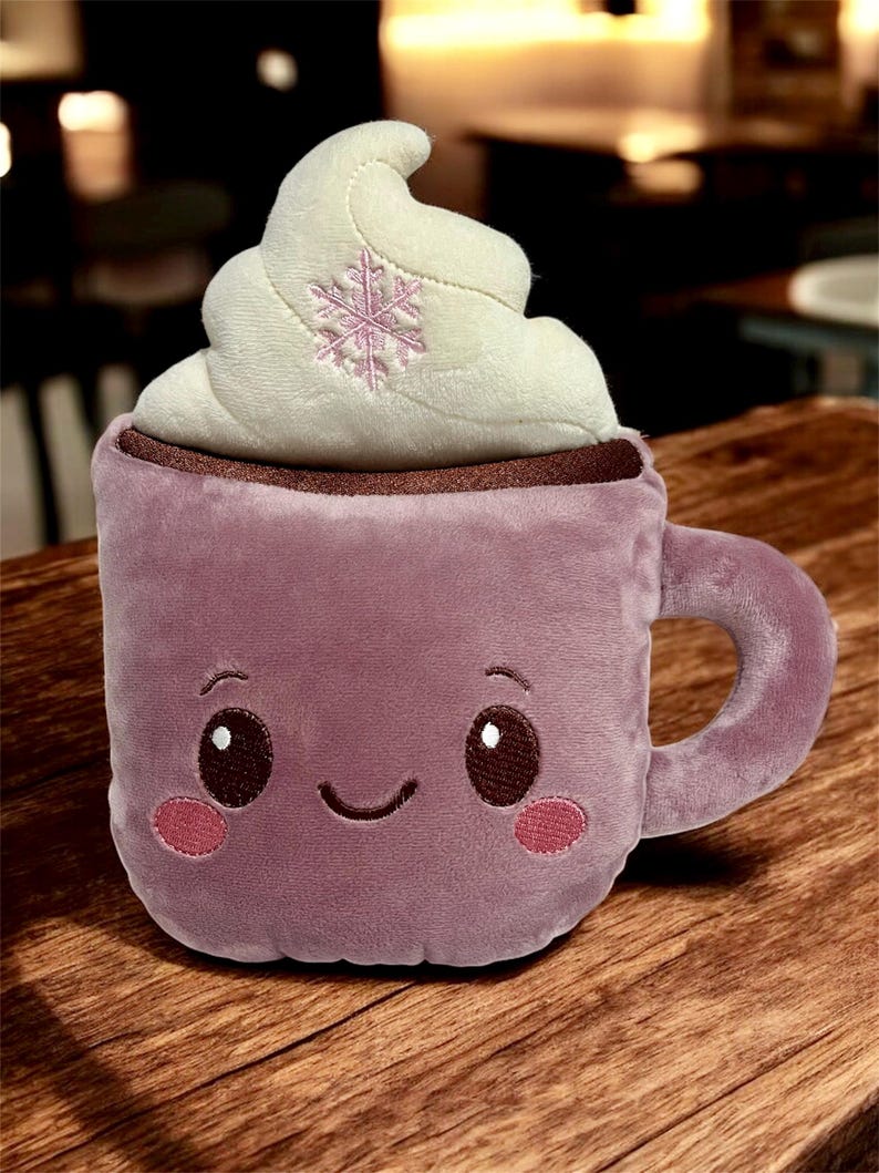 May include: Plush purple mug with a smiling face, pink cheeks, and a whipped cream topping. The whipped cream has a pink snowflake embroidered on it. The mug has a brown rim and a handle.