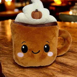 May include: A plush, brown coffee mug with a smiling face, topped with white whipped cream and a small pumpkin. The mug has a handle and pink blush marks on its cheeks.