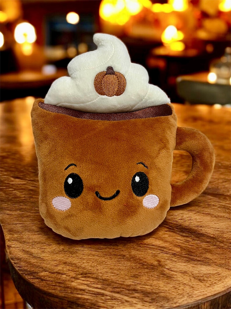 May include: A plush, brown coffee mug with a smiling face, topped with white whipped cream and a small pumpkin. The mug has a handle and pink blush marks on its cheeks.