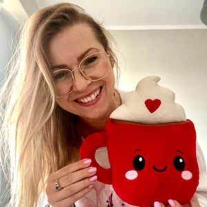 May include: A plush red mug with a smiling face, whipped cream topping, and a heart. The mug has a handle and is held by a person. The mug is a cute, whimsical design.