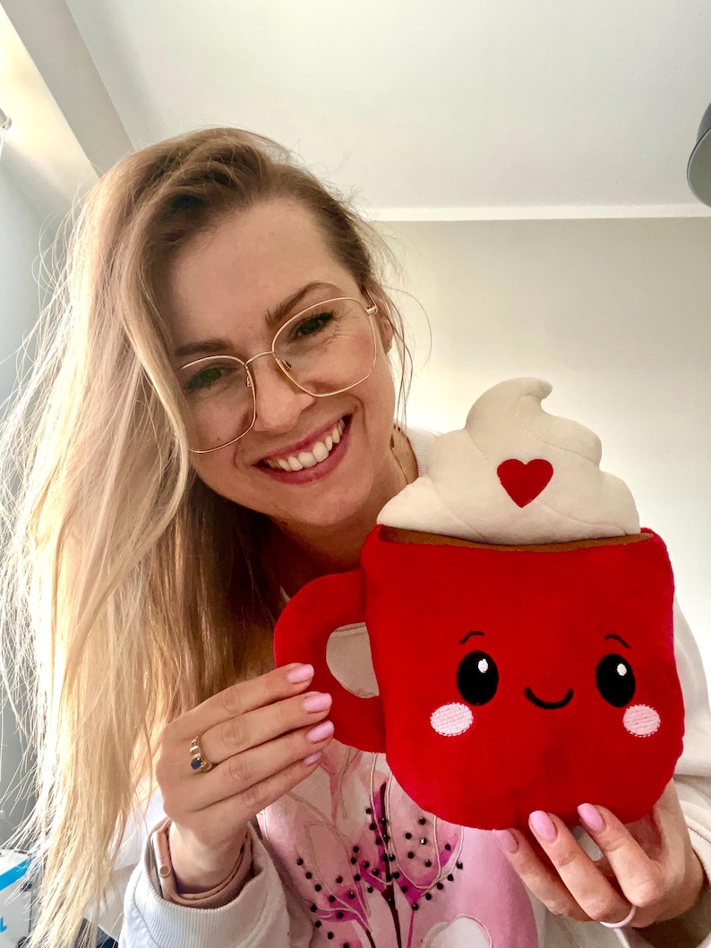May include: A plush red mug with a smiling face, whipped cream topping, and a heart. The mug has a handle and is held by a person. The mug is a cute, whimsical design.