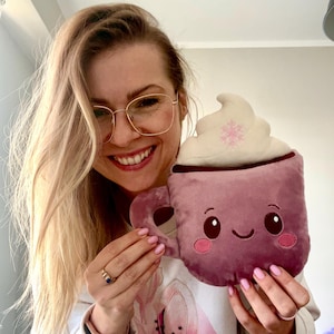 May include: Plush, mauve-colored mug with a whipped cream topping and a snowflake design. The mug has a smiling face and a handle. The plush toy is held up by a person.