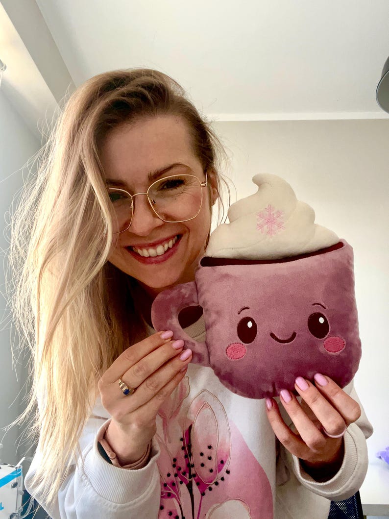 May include: Plush, mauve-colored mug with a whipped cream topping and a snowflake design. The mug has a smiling face and a handle. The plush toy is held up by a person.