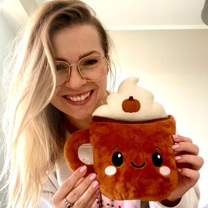 May include: A plush, brown coffee mug with a smiling face, white whipped cream, and a small pumpkin on top. The mug has a handle and is held by a person.