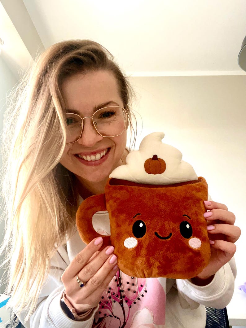 May include: A plush, brown coffee mug with a smiling face, white whipped cream, and a small pumpkin on top. The mug has a handle and is held by a person.