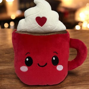 May include: A plush red mug with a smiling face, topped with white whipped cream and a red heart. The mug has a brown rim and a handle. The toy is designed to look like a hot chocolate.