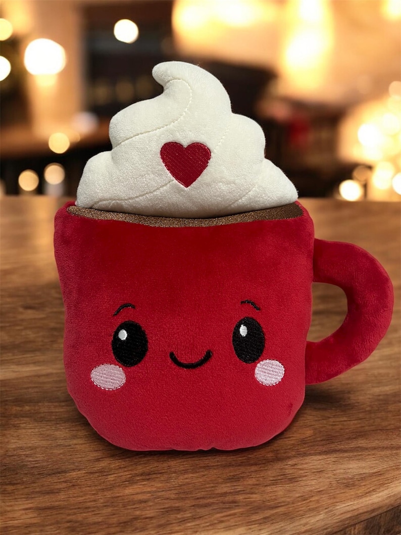 May include: A plush red mug with a smiling face, topped with white whipped cream and a red heart. The mug has a brown rim and a handle. The toy is designed to look like a hot chocolate.