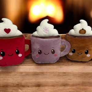 May include: Three plush hot chocolate mugs with whipped cream topping. The mugs are red, purple, and brown, each with a smiling face and a different design on the whipped cream: a heart, snowflake, and pumpkin.