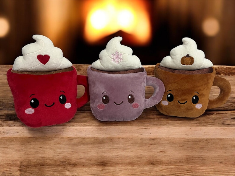May include: Three plush hot chocolate mugs with whipped cream topping. The mugs are red, purple, and brown, each with a smiling face and a different design on the whipped cream: a heart, snowflake, and pumpkin.
