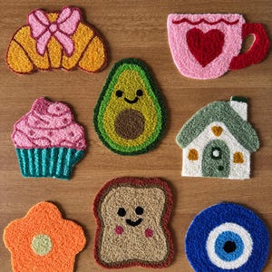Puede incluir: Una colección de posavasos coloridos y texturizados en varias formas. Los diseños incluyen un cruasán con un lazo rosa, un aguacate, una taza de té con un corazón, una magdalena, una casa, una flor, una rebanada de pan tostado y un ojo azul.