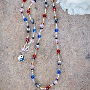 Puede incluir: Un collar de cuentas de colores con cuentas rojas, azules, rosas y transparentes. El collar tiene un cierre plateado y un pequeño colgante con el símbolo del yin-yang. El collar se muestra sobre una superficie texturizada de color marrón claro.