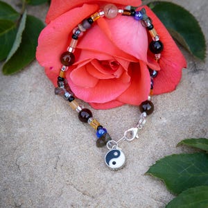 Puede incluir: Una pulsera de cuentas con un dije de Yin Yang, con una mezcla de cuentas marrones, negras y transparentes. La pulsera se muestra sobre una rosa rosa y una superficie de piedra.