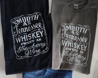 Può includere: Due magliette, una nera e una grigia, con la scritta "SMOOTH as Tennessee WHISKEY SWEET AS Strawberry Wine" in bianco. Le magliette sono piegate, con uno scorcio di denim blu.