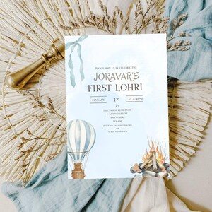 May include: A light blue invitation card for Joravar's First Lohri, featuring a watercolor hot air balloon with a teddy bear and a bonfire illustration. The card includes the date, time, and location details.