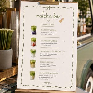 Puede incluir: Un letrero de menú blanco en un portapapeles de madera titulado matcha bar, con ilustraciones de cinco bebidas de matcha helado con descripciones para sabores de arándano, fresa, chocolate blanco, biscoff y pan de plátano, con un icono de corazón enjoy.