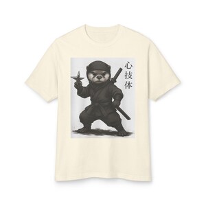 May include: Cream-colored t-shirt featuring a graphic of a ninja otter holding a shuriken and a sword. The otter is dressed in a black ninja outfit. Japanese characters are printed on the right side of the graphic.