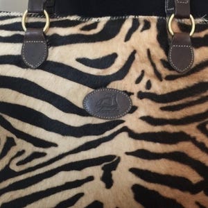 May include: A rectangular tote bag featuring a black and tan zebra-striped animal print pattern. The bag includes dark brown leather handles attached with gold-tone metal rings and a central oval leather patch embossed with a ship logo and text.