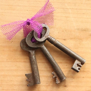 May include: Three antique metal keys tied together with a purple mesh ribbon. The keys have intricate designs and a rustic, aged appearance. The background is a light-colored wooden surface.