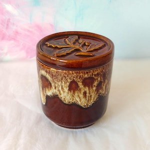 May include: A brown ceramic jar with a lid featuring a leaf design. The jar has a dark brown base with a lighter, dripped glaze effect towards the top. The lid is also brown.
