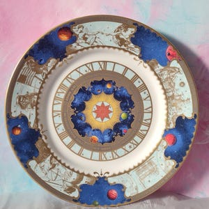 May include: A decorative plate with a celestial theme. The plate has a white centre with a detailed illustration of a sun, planets, and stars. The rim is adorned with Roman numerals, celestial figures, and a gold border. The plate's design includes blue, gold, red, and yellow.