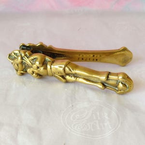 May include: A gold-coloured brass nutcracker shaped like a figure with articulated arms and legs. The metal surface features engraved details and the text "ENGLAND" stamped on the inner side of the handle, showing a metallic finish and texture.