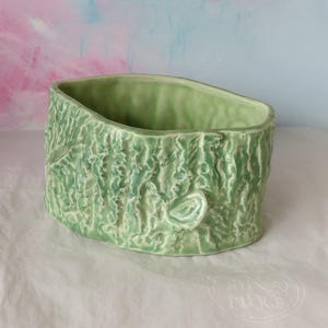 May include: A pale green ceramic planter with a textured, bark-like exterior. The oval-shaped container has a smooth interior and a naturalistic design, suitable for small plants or decorative use. The background is a soft blend of pink and blue.