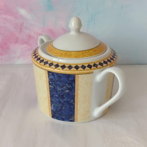 May include: A ceramic sugar bowl with a lid, decorated with a yellow and blue geometric pattern. The bowl has two handles and a lid with a small knob. The design includes a blue vertical stripe and a yellow and blue diamond pattern.