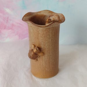 May include: A tan ceramic vase with a wavy rim and a cylindrical body. Two small turtle figurines are attached to the vase, one on the side and one near the rim. The vase is approximately 18 cm tall.
