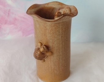 May include: A tan ceramic vase with a wavy rim and a cylindrical body. Two small turtle figurines are attached to the vase, one on the side and one near the rim. The vase is approximately 18 cm tall.