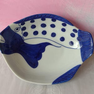 May include: A ceramic fish-shaped dish, white with blue painted details. The fish has a smiling face, dotted pattern, and curved fins. The dish is designed for serving food.