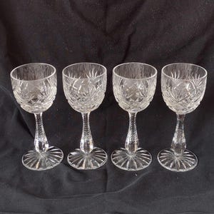 May include: Four clear crystal wine glasses with detailed starburst and diamond patterns. Each glass has a round base, a detailed stem, and a bowl-shaped top. The glasses are arranged in a row against a dark background.